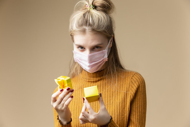 Blond Female Girl with Tiny Small Yellow Gift Christmas Box Wearing Facial Protective Mask Against Viruses and Unwrapping Present