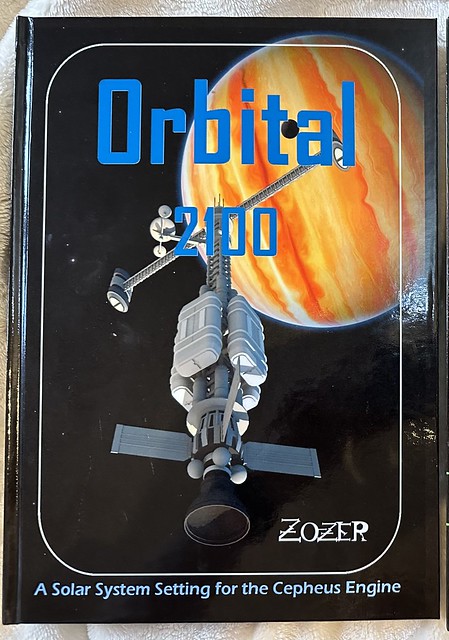 The cover of Orbital 2100, which shows an in-system spacecraft (with engines orientated to the bottom of the page) approaching Jupiter. The header is Orbital 2100 in Blue, and at the bottom of the page the sub-title says ‘A Solar System Setting for the Cepheus Engine’. The image is surrounded with an overlaid white rounded rectangle box.