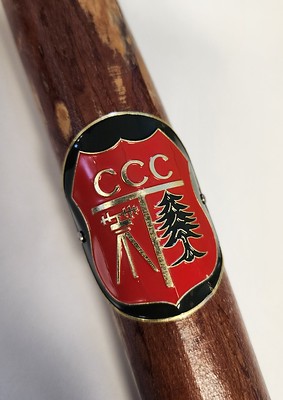 White surface with a zoomed up image of a wooden walking stick with a red CCC medal