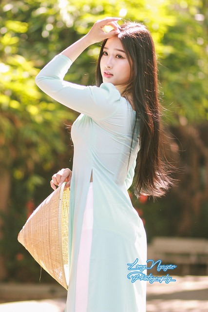 In the radiant glow of bright sunlight, a beautiful girl in a resplendent light blue Traditional Vietnamese Dress and a Vietnam Bamboo Hat was posing gracefully in the front yard of the communal house, where red flowers bloom in vibrant splendor.