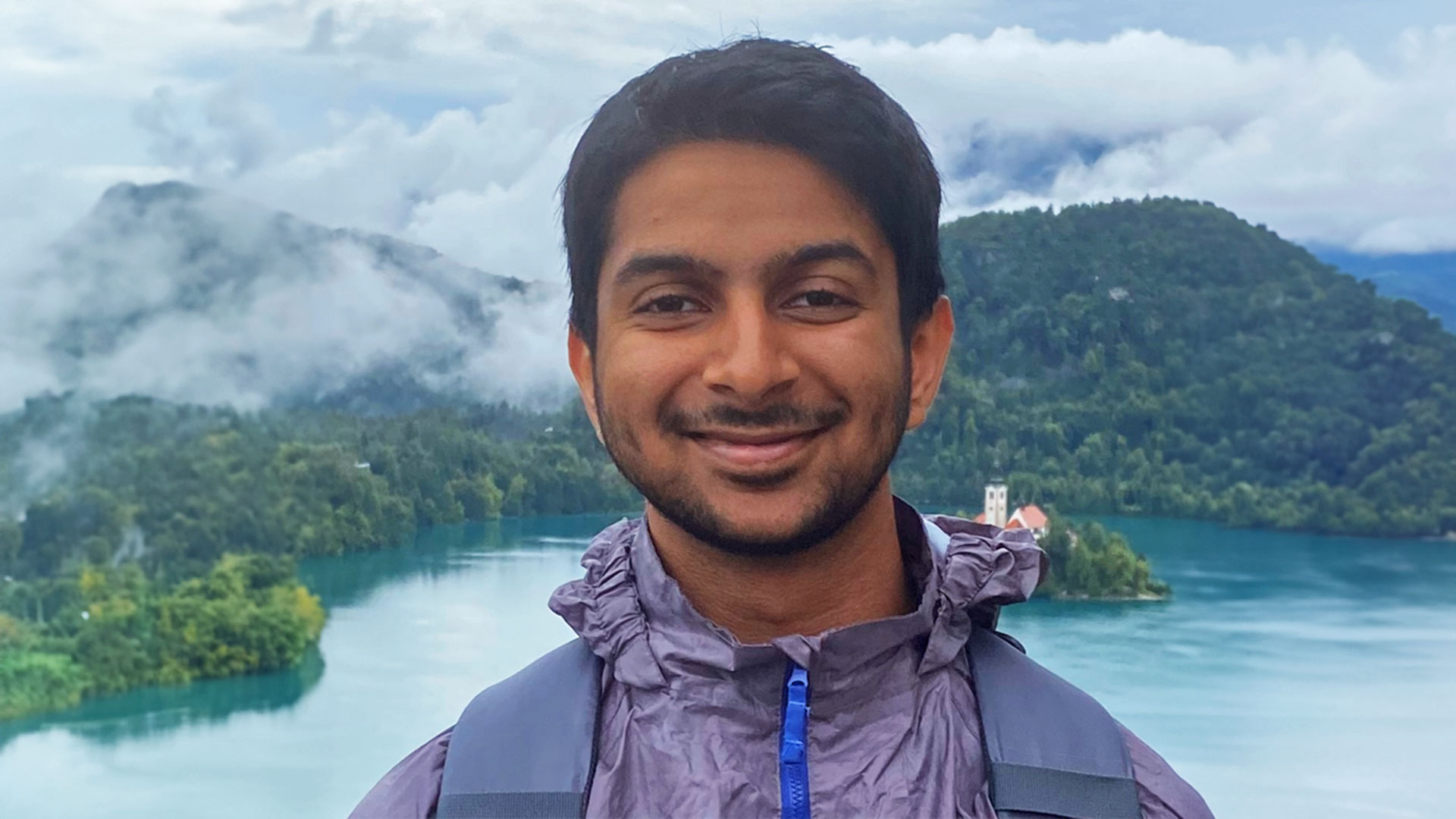Anand smiling to the camera in front of a backdrop of a lake.