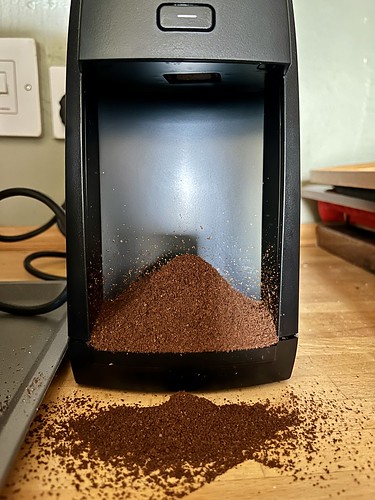 A photo of a pile of ground coffee that's come out of the bottom of a coffee grinder, not caught by anything, spilling on to a wood counter