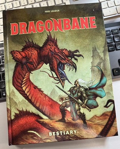 Picture of a book lying on a grey desk, with two keyboards underneath it, a black one in the middle of the screen and a white Apple one to the top above it. The book has an image of a knight with flowing white long hair and a blue-green cloak facing down a red coloured wingless dragon (perhaps a Lindworm) which rears above him as it's tail wraps around the base of the book. A castle looms out of the mist in the background on the right. The top of the book says "Free League" and has the red Dragonbane logo, and the bottom of the book says "Bestiary".
