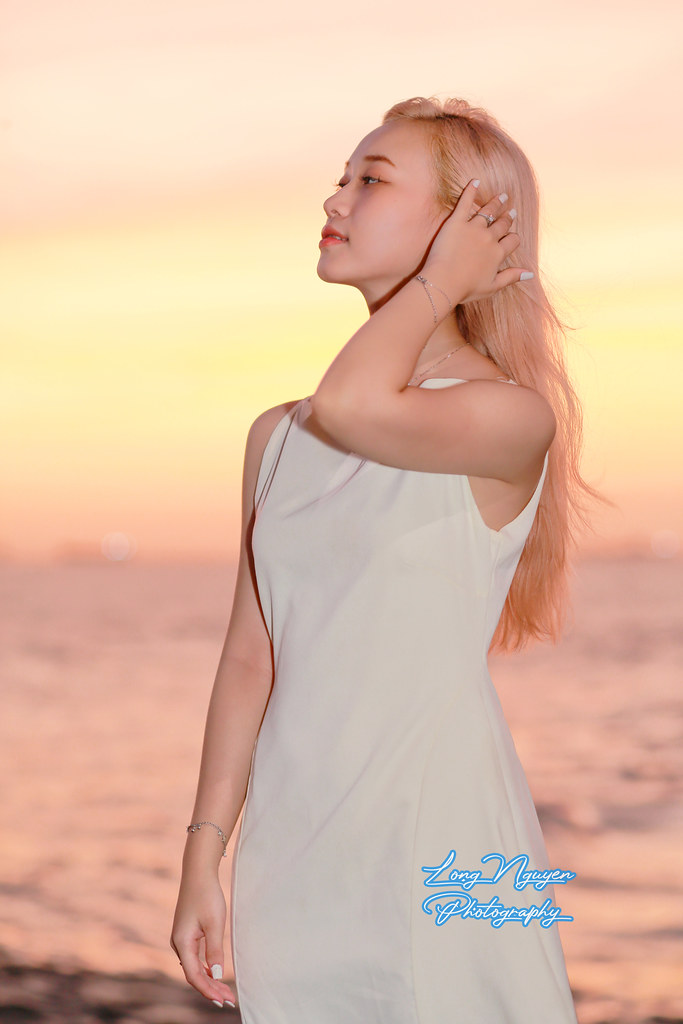 In the coastal sky's brilliance, a captivating girl strikes a pose on a cement platform, beneath the hues of a purple-pink sunset descending over the sea.