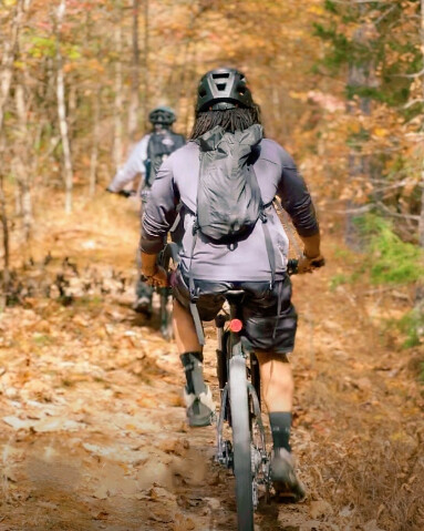 A Black man on a mountain bike faces away from the camera, riding his bike on a mountain biking trail with ample fall foliage surrounding him.