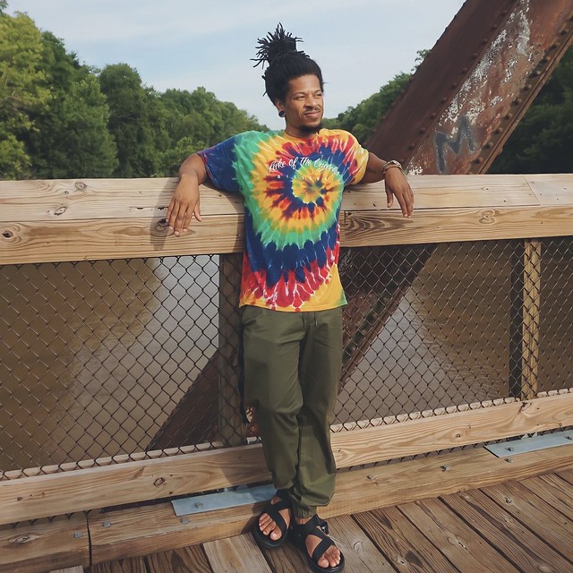 A Black man wearing a colorful tye-dye shirt leans on the rails of Staunton River Bridge smiling. The bridge is over a brown river with green trees in the background.