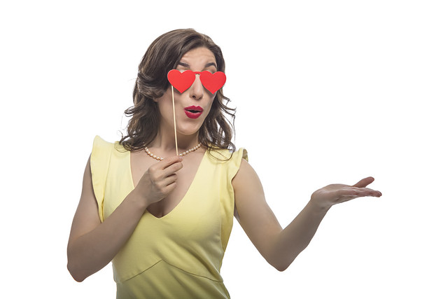 Cute and Funny Positive Smiling Caucasian Brunette Woman With Red Heart Shaped Artistic Spectacles In Front Of Face Over White Hiolding hand In Front