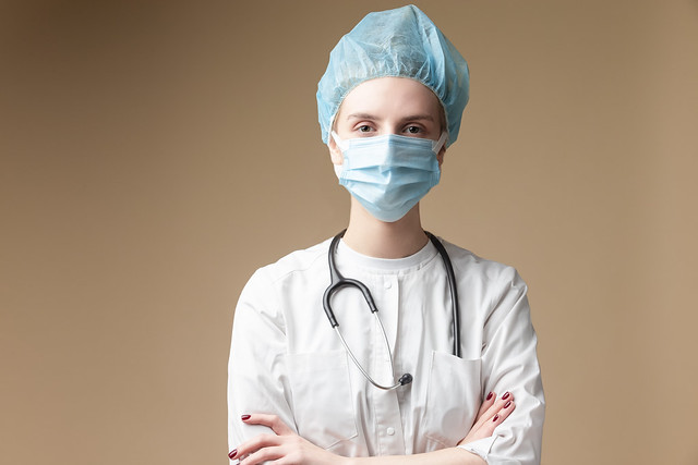 Professional Female GP Doctor Posing in Doctor's Smock and Facial Mask and Endoscope With Hands Folded Over Beige.