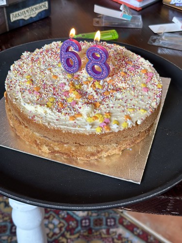 A white sponge cake with white icing covered in sprinkles and two candles making '68'.