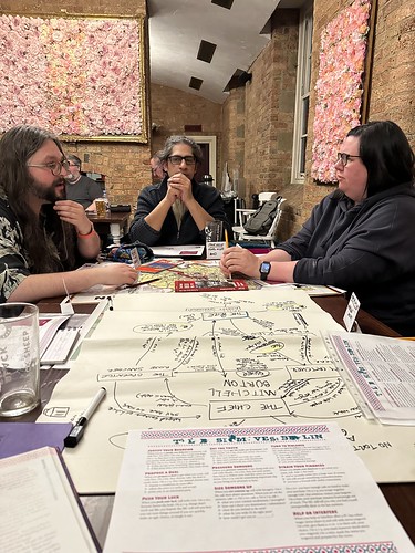 A picture mid-session at a gaming table at Revelation. Two players discuss the potential fate of another player's character, while that player pensively looks on. The foreground has move sheets and the relationship map (inverted from the POV). A map of Cold War Berlin and guidebooks sit between the players.