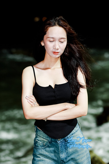 A captivating maiden, with lustrous black and red-tinted locks, stands on concrete steps beside a rocky beach. Bathed in soft electric light against the dark, undulating waves with frothy white foam, she strikes a graceful pose.