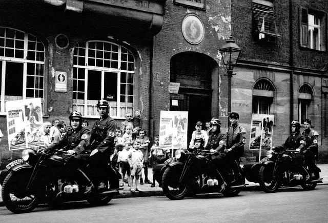 Signs advertise a motorcycle race behind members of the NSKK in Doos-Nürnberg, Germany 1937