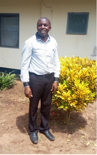 Pius Kilasy in white shirt and black pants in front of yellow bush in Tanzania.
