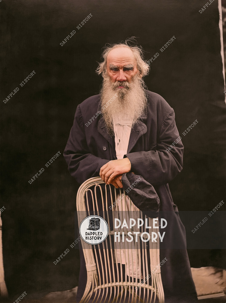 Portrait of Tolstoy, in a black coat over a white blouse. He stands, leaning his folded hands on the back of a wicker chair, holding a black cap in his right hand. Year: 1908. Place: Yasnaya Polyana, Tula Oblast, Russia. Photographer: Karl Bulla.