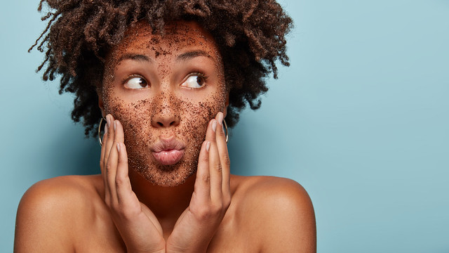 Curious black lady focused aside has coffe scrub on face applies mask on face, focused aside, has bare shoulders, isolated over blue background with blank space for promotion. Body care. Cosmetology