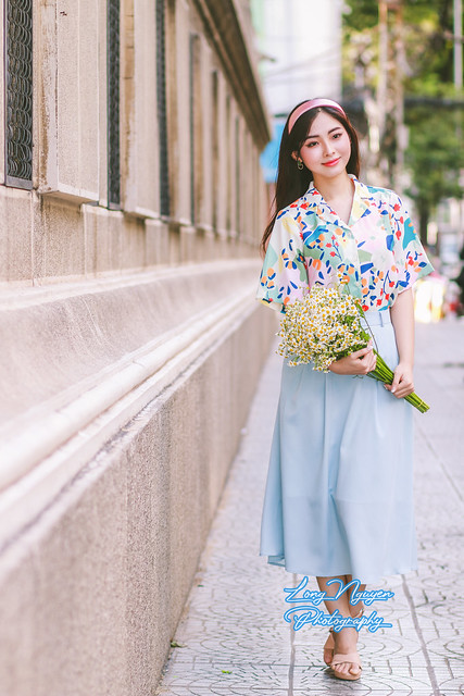 On a street corner, a lovely maiden dons a turban, its fabric intertwined with her yellow-black hair. Wrapped in a Jaryl Floral Wrap shirt and a light blue skirt, she holds a bouquet of white daisies, exuding happiness and confidence in her pose.