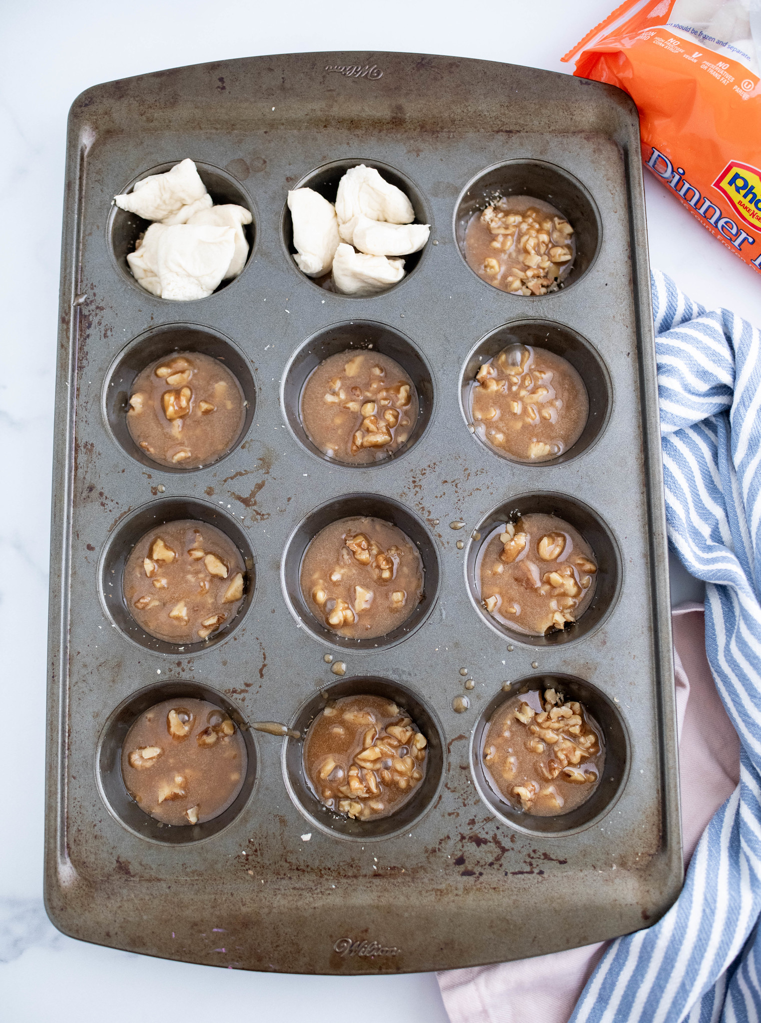 Muffin pan with pecans and caramel in each muffin cup. Pieces of bread dough in two of the muffin cups.