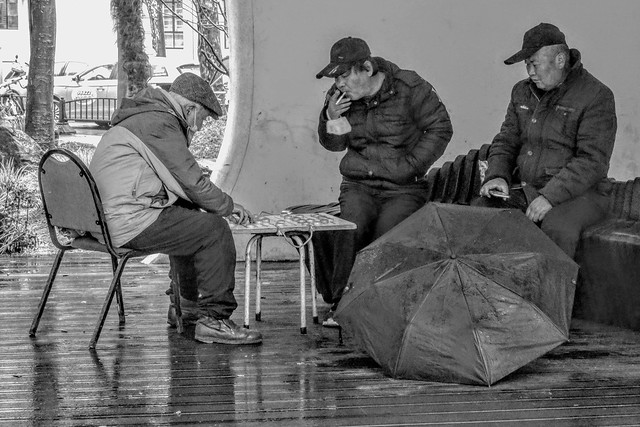 Elderly men playing Chinese chess in a pavilion on a green space in the street on a cold and rainy day