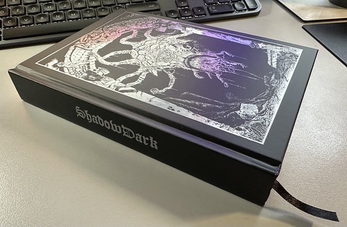 A black and silver digest sized hardback book lies on a light-grey desk in front of a black and dark-grey keyboard. The cover of the book has no text, just a horrific creature floating in an archway, all teeth and tentacles with orbs or eyes at the end. The spine of the book has the word 'Shadowdark' in a gothic style.