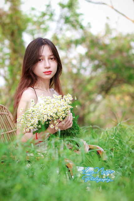 On the expansive green lawn, a lovely maiden dons a pristine white dress, her silky red-blonde hair cascading. Seated on a rattan chair, she holds a bouquet of White Daisies, posing at angles that reveal varied backgrounds aglow in the afternoon sunlight.
