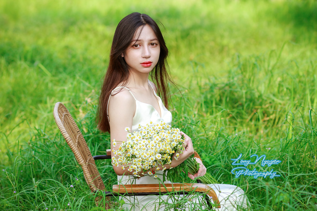 On the expansive green lawn, a lovely maiden dons a pristine white dress, her silky red-blonde hair cascading. Seated on a rattan chair, she holds a bouquet of White Daisies, posing at angles that reveal varied backgrounds aglow in the afternoon sunlight.