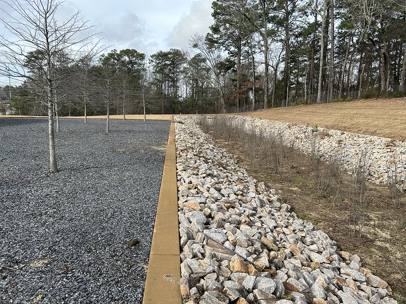 A rectangular area with rocks is quiet and dry and grey, next to a seating area with a few dormant trees.