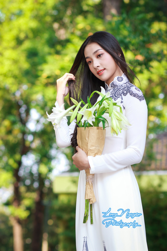 In a pristine white traditional Vietnamese dress adorned with simple patterns, a lovely maiden holds a bouquet of white lilies. Shiny black hair cascades over her shoulders as she poses brilliantly beneath resplendent foliage in the afternoon sunlight.