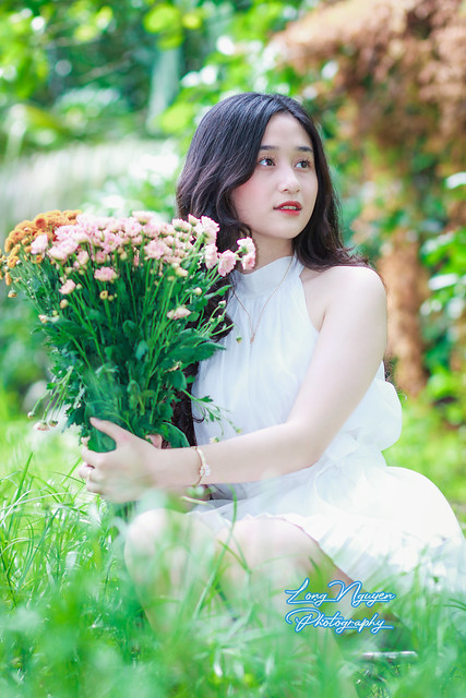 A lovely muse, clad in a pristine white gown with flowing black hair, sits upon a chair amidst the verdant lawn. With a cheerful demeanor, she poses gracefully, holding a bouquet of flowers in the morning light.