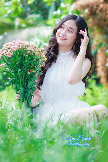 A lovely muse, clad in a pristine white gown with flowing black hair, sits upon a chair amidst the verdant lawn. With a cheerful demeanor, she poses gracefully, holding a bouquet of flowers in the morning light.