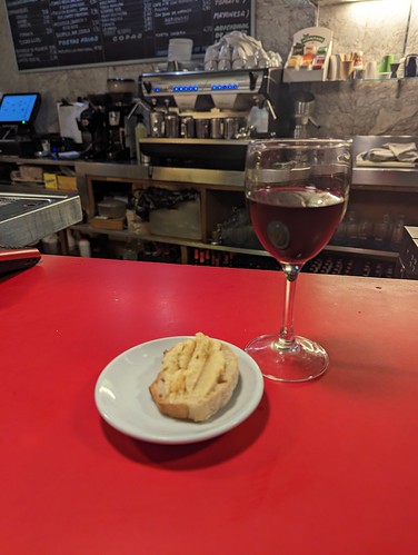 A glass of wine on a red bar top with a small plate containing a piece of bread with a spread on top
