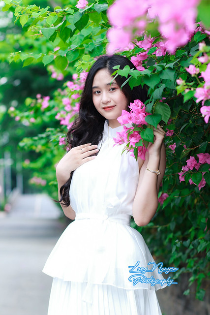 A lovely muse in a pristine white gown, her silky black curls cascading over her shoulders. She stood joyfully and innocently on the roadside, beside a wall adorned with vibrant pink bougainvillea branches in morning sunlight.