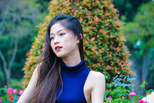 A lovely young girl, dressed in a short sweater and blue jeans, strikes a confident pose, her radiant face expressing charm. The backdrop features morning sunlight casting a warm glow on lush green trees.