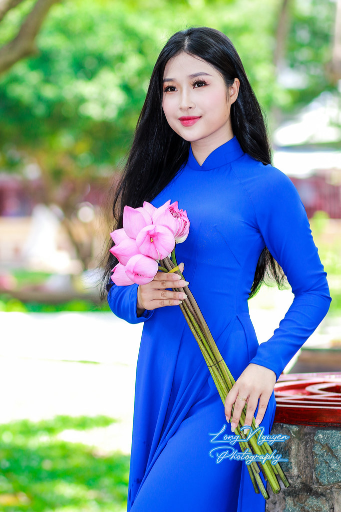 In the serene morning light, a lovely maiden, her delicate face framed by straight, silky black hair, stands by an ancient well and banyan tree. Clad in a blue Vietnamese dress, she holds a bouquet of pink lotus flowers, exuding innocence and cheer.