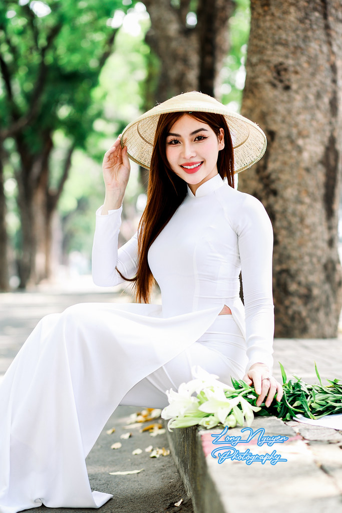 An elegant girl in a white traditional Vietnamese dress and Asian conical hat sits gracefully on a park step amidst towering old trees. Beside her rests a bouquet of lilies, illuminated by the morning sunlight filtering through the lush green foliage.