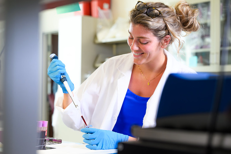 A smiling woman uses a pipette to transfer blood to a test tube.