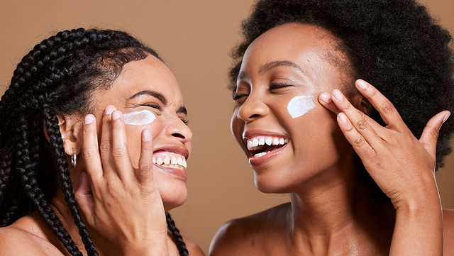 Black woman, cream and skincare with friends support, love and beauty together in studio for cosmetics, makeup cleaning and facial. Excited, diversity model with sunscreen or essential oil on face