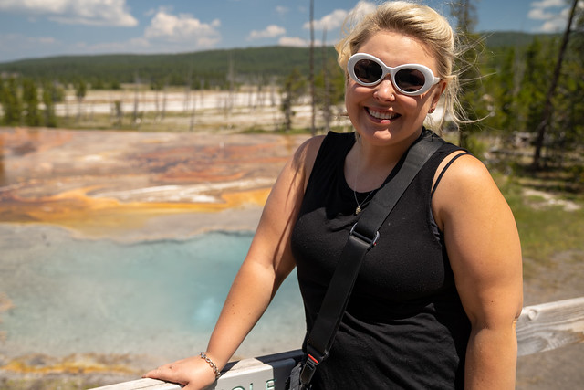 Pretty blond woman tourist poses in front of Firehole Spring in Yellowstone National Park, wearing white sunglasses