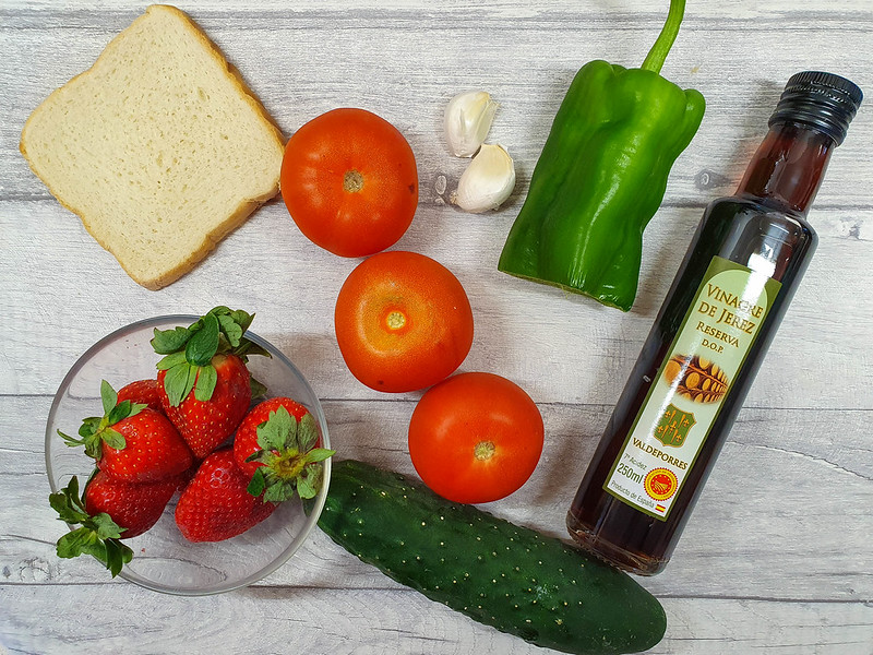 Some of the ingredients for the Spanish gazpacho recipe: three tomatoes, a cucumber, half a green bell pepper, a bottle of Jerez vinegar, a glass bowl with strawberries, two garlic cloves and a slice of stale bread.