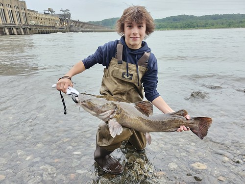 Photo of boy in the water near a dam, holding a fish. 