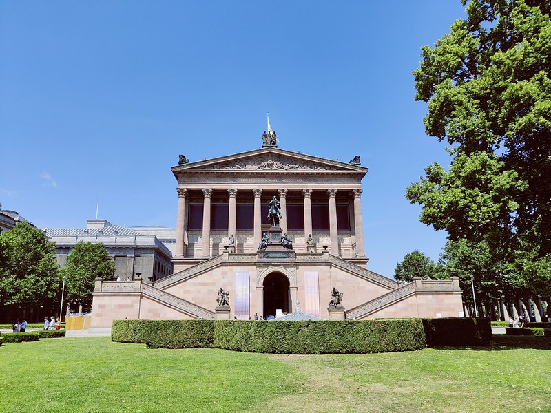 A museum looking like a Roman temple, on the Museum island. It has stairs leading to it, from a beautiful green lawn. The sky is blue without any clouds.