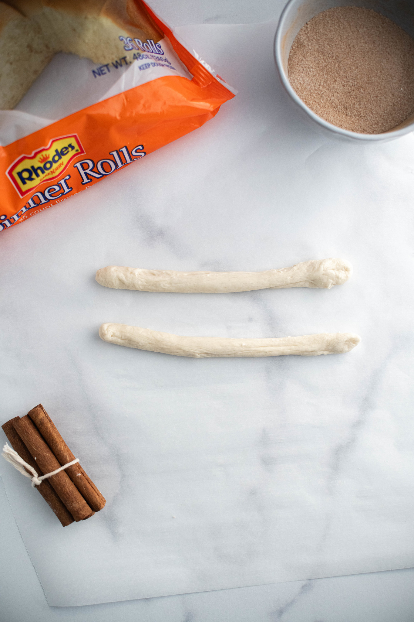 Two rope of dough rolled out on parchment with a small bowl of cinnamon sugar and package of Rhodes rolls off to the side.