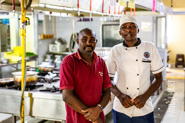 Two men stand in a bright commercial kitchen; left in a red polo, right in a white chef's jacket and hat, with stainless steel counters and cooking pots behind them.