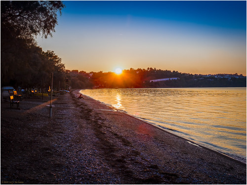 Sunset Reflections on Paralia Pigadakia Chalkoutsi's Serene Shore by Luc V. de Zeeuw, Warm sunlight dips below the horizon, casting golden hues on Paralia Pigadakia Chalkoutsi's tranquil