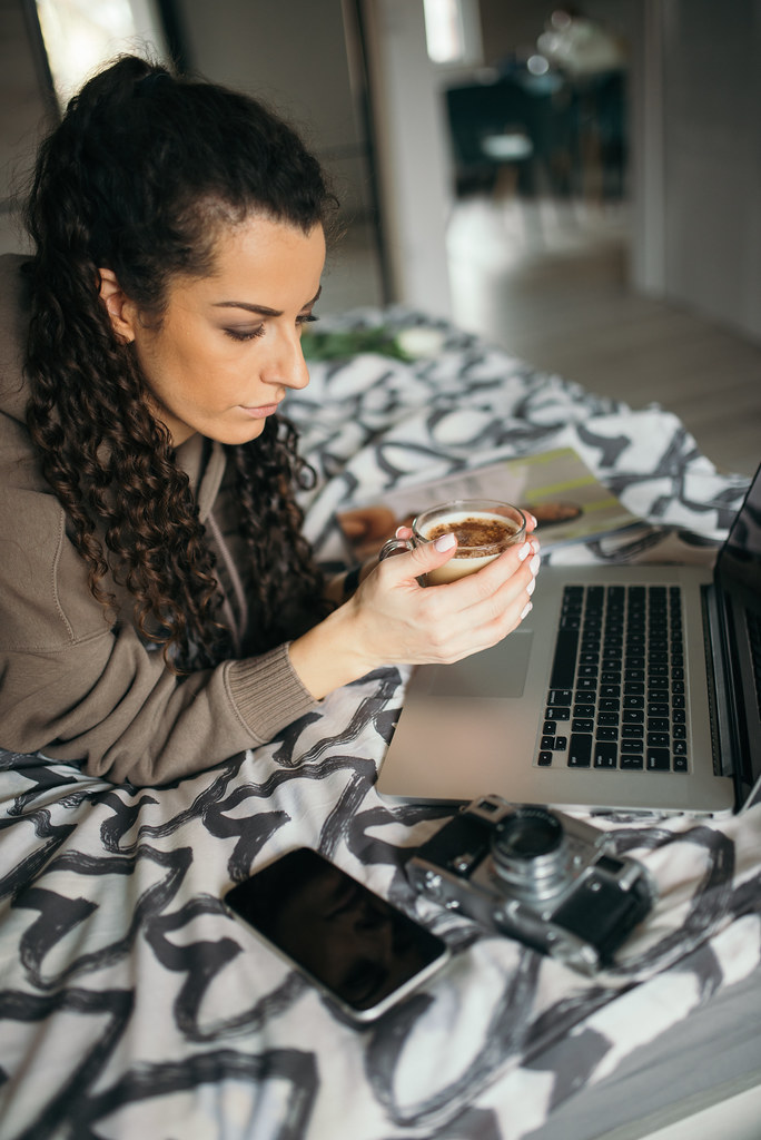Woman lying in bed and holding a cup of coffee while looking at her laptop.