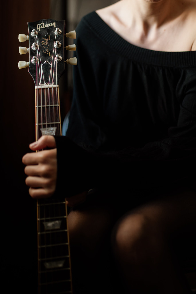 A person in a black off-shoulder top holds a guitar close, exuding a relaxed vibe in a warm indoor environment
