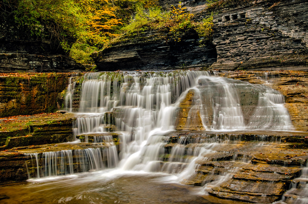 A wide cascade steps down stone ledges below trees turning orange and yellow along the gorge wall.