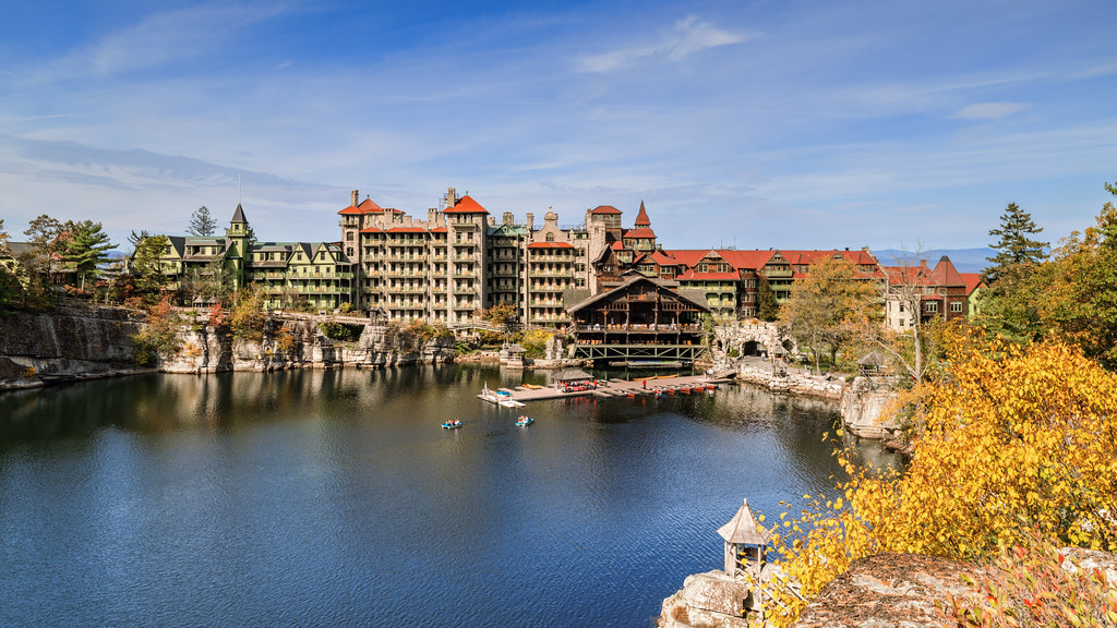 The long hotel rises over Mohonk Lake with yellow and orange shrubs and trees on the cliff edge in front and a few turning crowns along the shore.