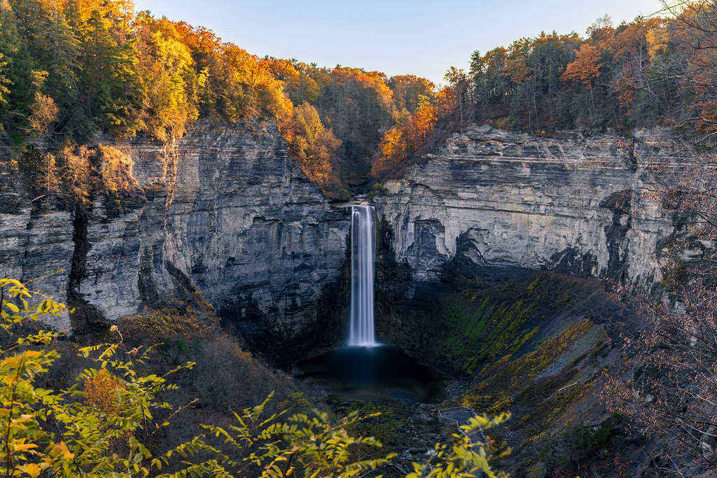 Evening sun lights orange and gold oaks along the gorge rim above the falls, with bare trees on the slope below.