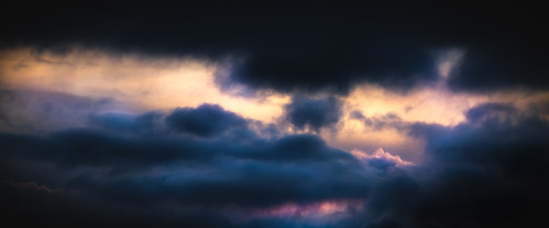 "A moody sky filled with dark, dense clouds, broken by glowing streaks of orange, pink, and purple light from the setting sun, creating a dramatic contrast of shadow and illumination." | by ScruffsClubPhotography