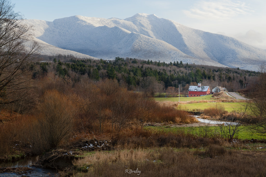 Bare trees and brown brush line a stream before a red barn, with Mount Mansfield white with snow behind.